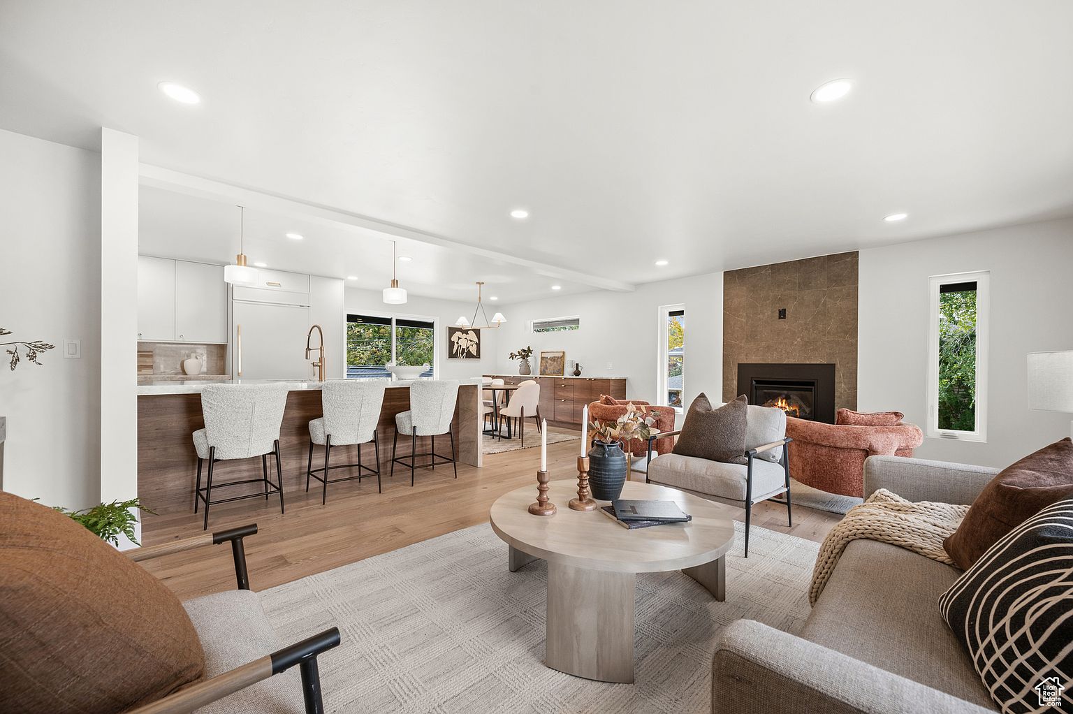 This is an interior shot of a living room that seamlessly blends into a kitchen and dining area. The living room features a fireplace with a stone surround, comfortable seating arrangements, and a neutral color palette. The open floor plan creates a spacious and inviting atmosphere, perfect for modern living.