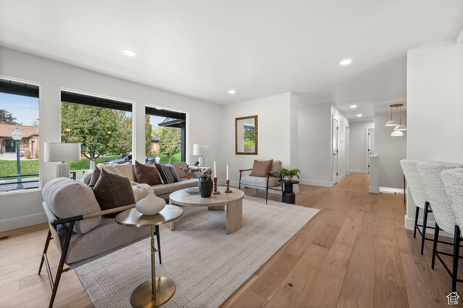 This is an interior shot of a living room featuring a neutral color palette with hardwood floors and a light-colored area rug. The room is furnished with a sofa, accent chairs, and a round coffee table, complemented by decorative items such as vases and candles. Large windows provide natural light and views of the outdoors, creating a bright and inviting atmosphere.