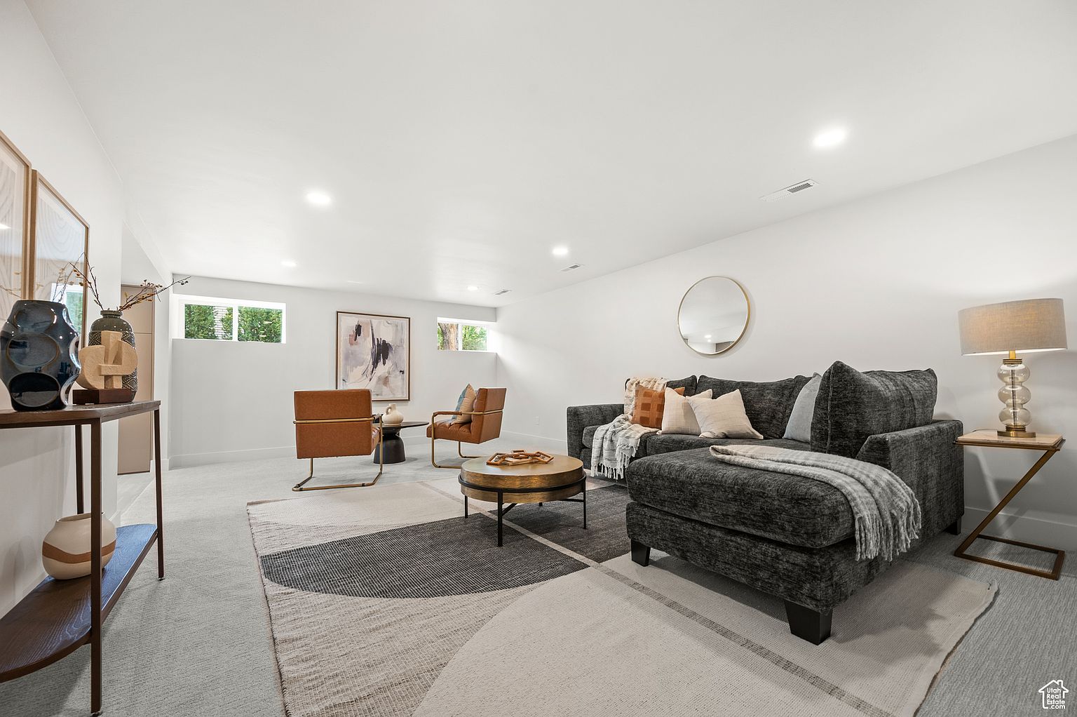 This is a well-staged living room featuring a gray sectional sofa with decorative pillows and a throw blanket, a round coffee table, and two leather armchairs. The room is decorated with a round mirror, artwork, and a side table with a lamp. The space is bright and inviting, with a neutral color palette and a modern aesthetic.