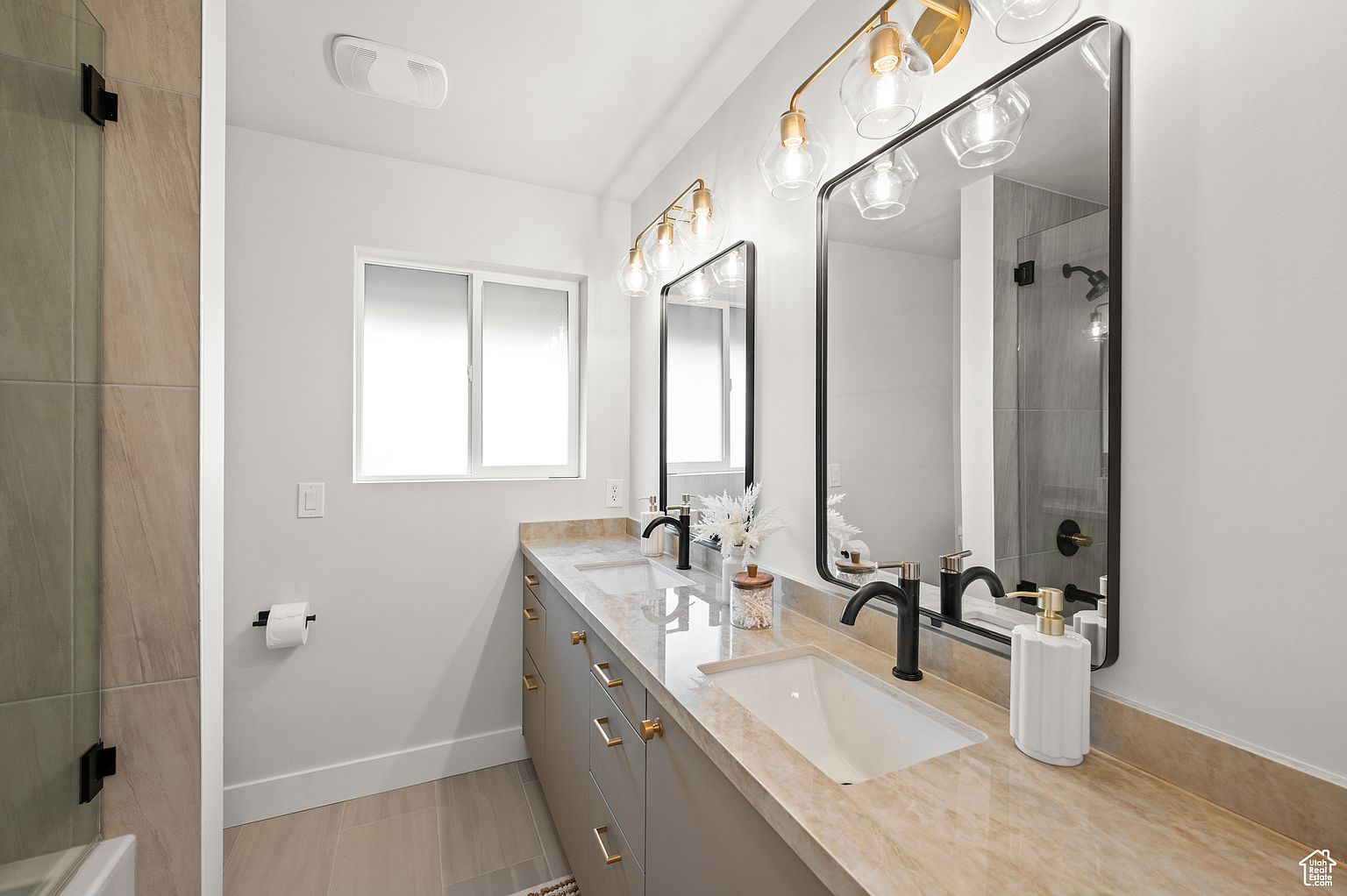 This is a well-lit primary bathroom featuring a double vanity with a light beige countertop and gray cabinets with gold hardware. Two black framed mirrors hang above each sink, illuminated by modern gold light fixtures. The bathroom has a clean and contemporary design with neutral color tones.