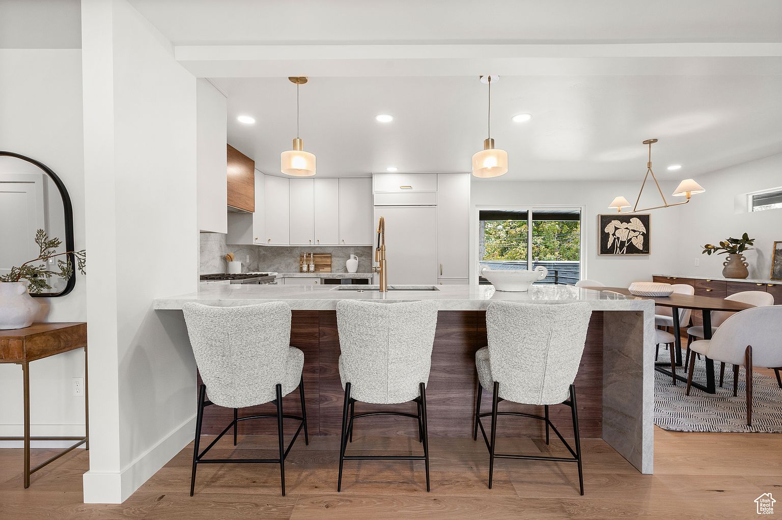 This interior shot showcases a modern kitchen with a breakfast bar featuring three stylish chairs. The kitchen boasts white cabinetry, marble countertops, and pendant lighting, creating a bright and inviting atmosphere. The open layout connects seamlessly to the dining area, enhancing the home's flow and functionality.