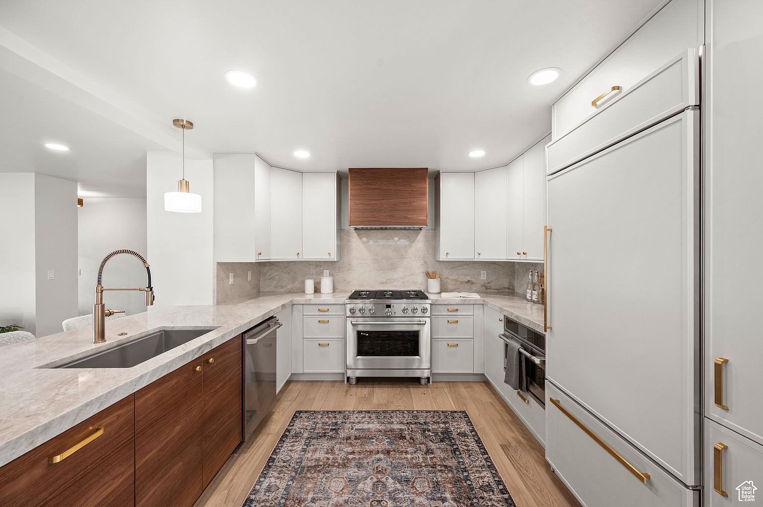 This is a well-lit kitchen featuring white and wood cabinetry, stainless steel appliances, and a marble countertop. The kitchen has a modern design with gold hardware and a stylish rug. The perspective is from the center of the kitchen, showcasing the layout and design elements.
