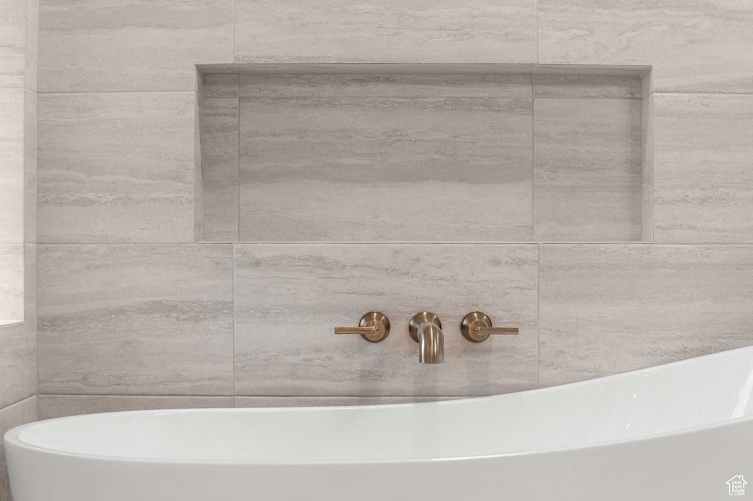 This image showcases a modern primary bathroom featuring a sleek, white freestanding bathtub with elegant bronze fixtures. The wall is adorned with large, horizontally-laid light gray tiles, complemented by a recessed niche for storage. The overall impression is one of understated luxury and spa-like tranquility.