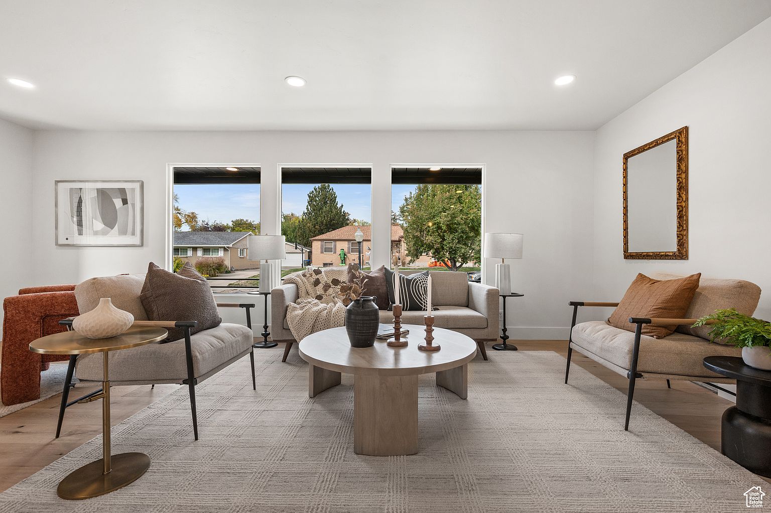 This is a bright and airy living room featuring a neutral color palette with pops of brown and black. The room is furnished with two armchairs, a sofa, and a round coffee table, all arranged on a patterned area rug. Large windows provide ample natural light and views of the outdoors, enhancing the inviting and comfortable atmosphere.