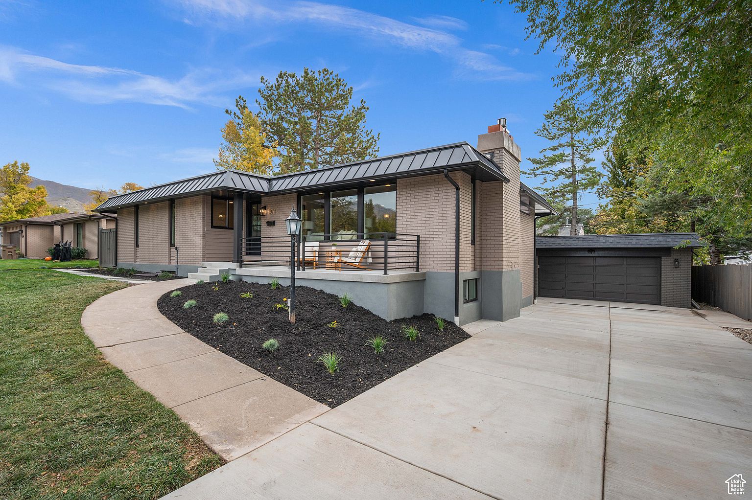 This is a front exterior view of a single-story brick home with a modern design. The house features a dark metal roof, a small front porch with a black railing, and a well-maintained lawn with a curved walkway leading to the entrance. A detached garage is visible on the right side of the house, and the landscaping includes dark mulch and small plants.