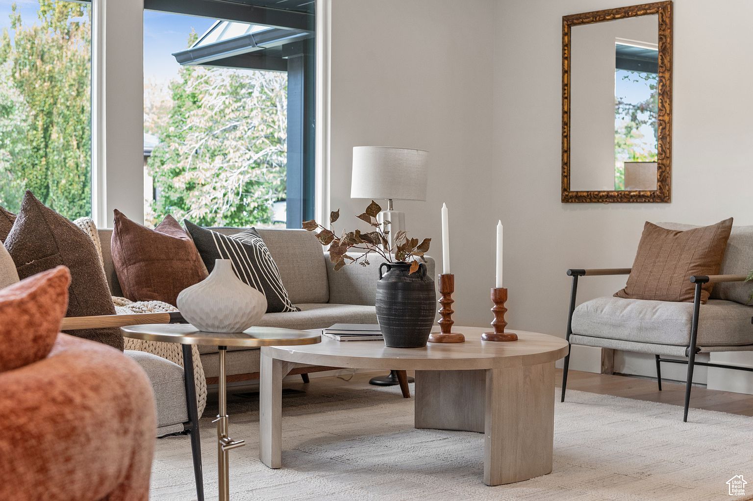 This is a well-lit living room featuring a neutral color palette with pops of brown and black. A comfortable sofa is adorned with decorative pillows, and a round coffee table displays vases and candles. The room is furnished with a chair and a side table, and a large window provides natural light and a view of the outdoors.