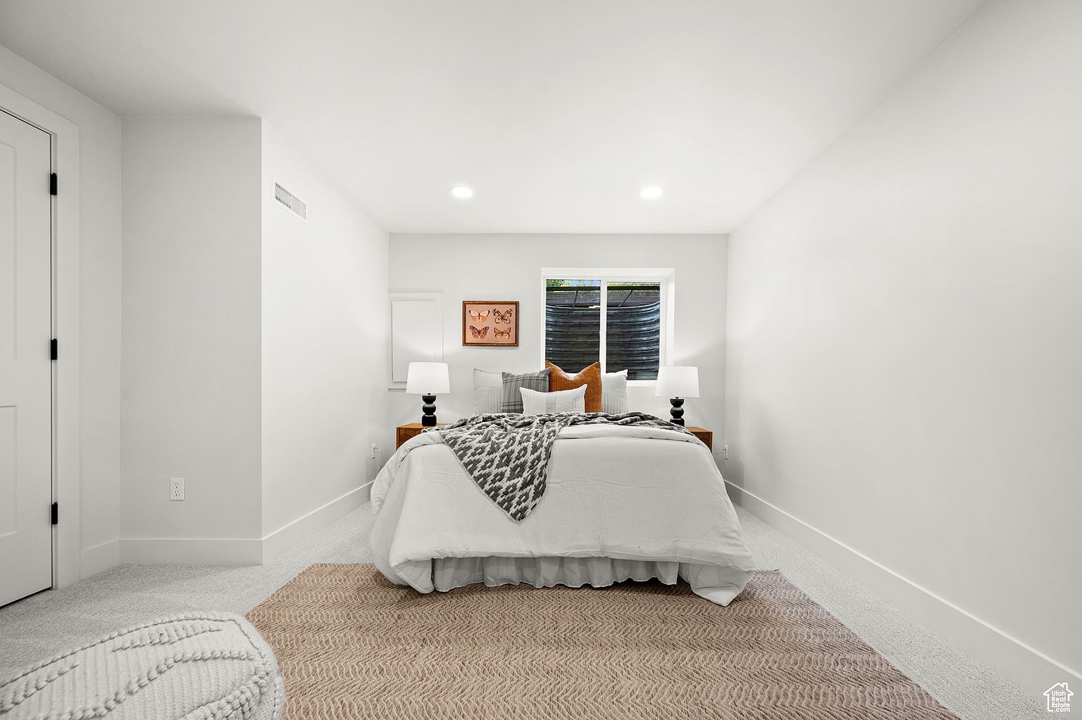 This is a bright and clean bedroom featuring a bed with white linens and a gray patterned throw. The room is painted white, with a neutral-toned rug adding warmth. A window provides natural light, and bedside lamps offer additional illumination, creating a cozy and inviting atmosphere.