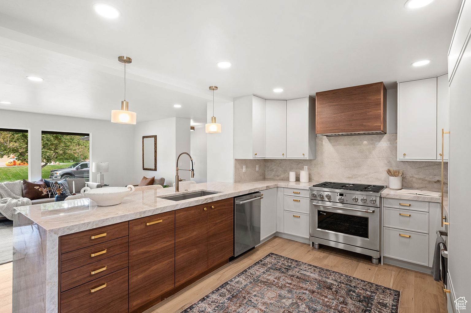 This is a bright and modern kitchen featuring a mix of white and wood-toned cabinetry, stainless steel appliances, and a large island with a light-colored countertop. The kitchen is well-lit with pendant lights and recessed lighting, and it opens to a living area, creating an open-concept space. A stylish rug adds warmth to the hardwood flooring.
