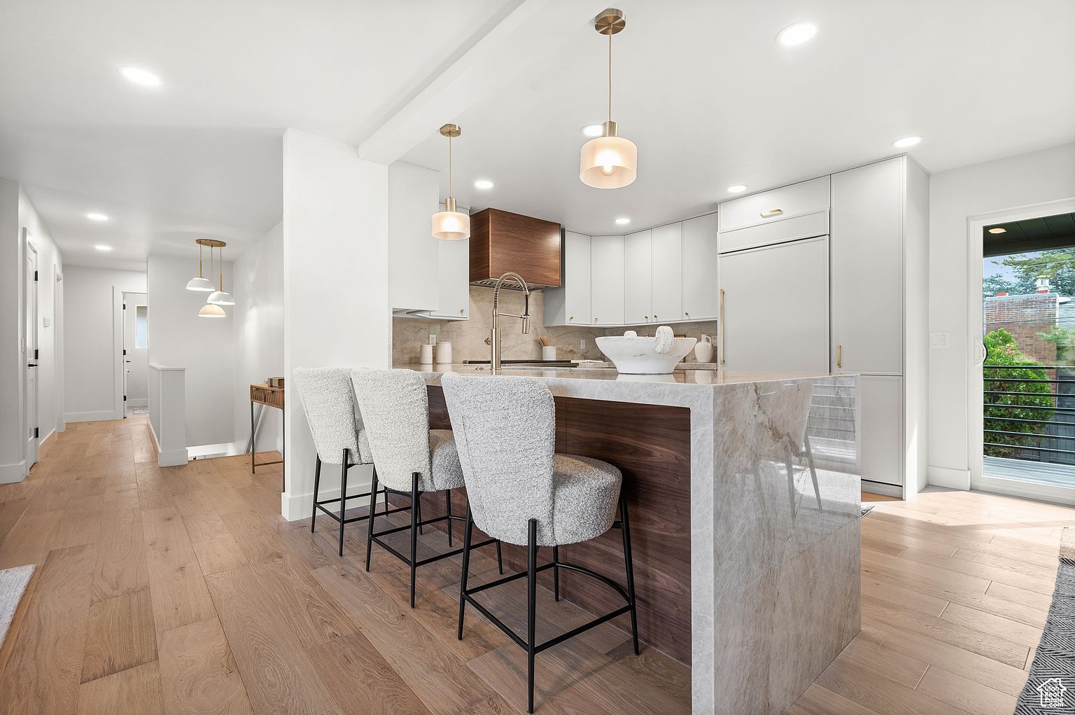This is a bright and modern kitchen featuring a large island with a marble countertop and wood accents, complemented by three stylish bar stools. The kitchen is equipped with white cabinetry and stainless steel appliances, illuminated by pendant lighting. The hardwood flooring adds warmth to the space, and a sliding glass door provides access to the outside.
