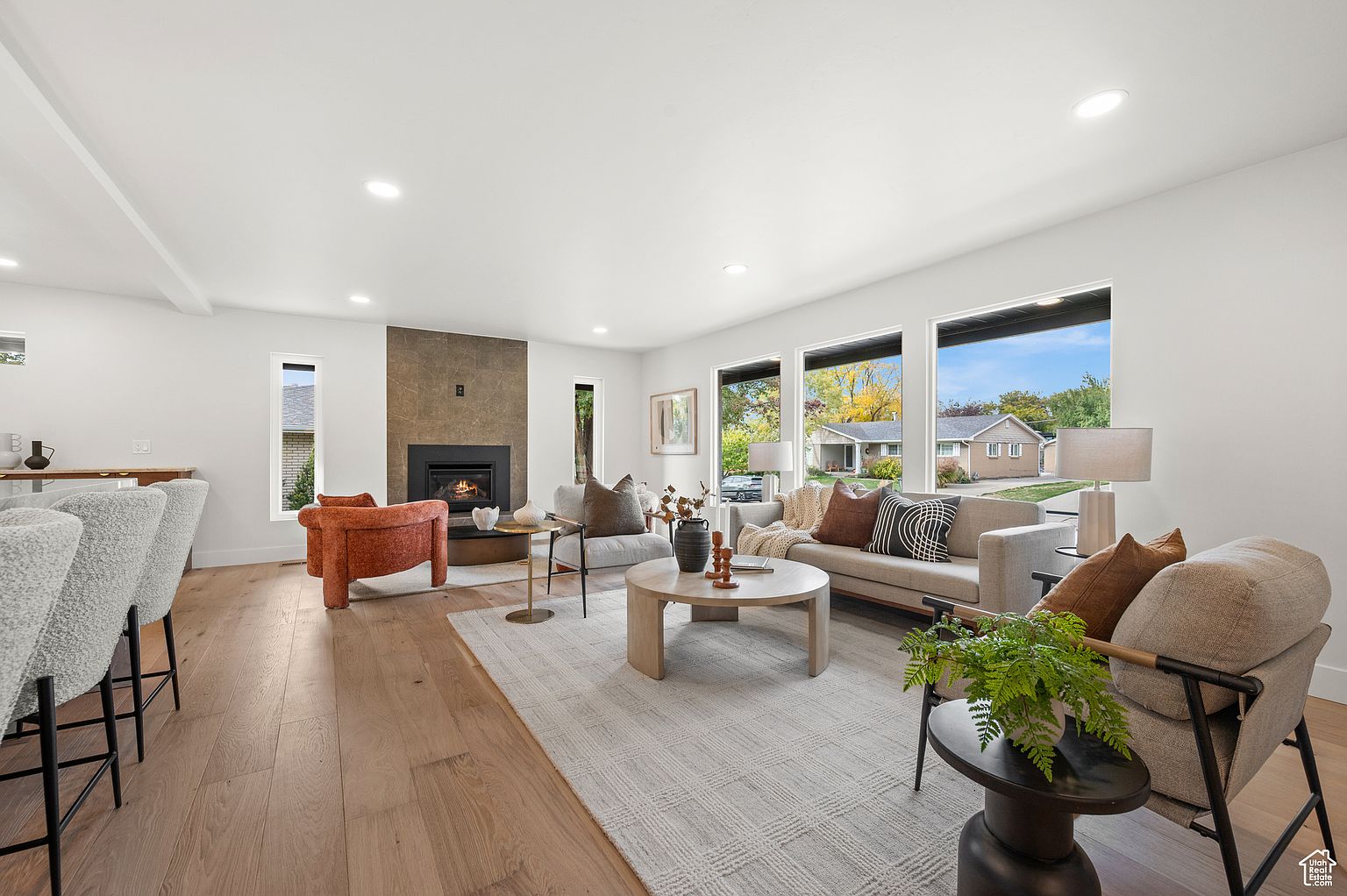 This is a bright and spacious living room featuring hardwood floors, white walls, and large windows that provide ample natural light. The room is furnished with a neutral-toned sofa, armchairs, a round coffee table, and a fireplace with a stone surround. A patterned area rug anchors the seating arrangement, creating a cozy and inviting atmosphere.
