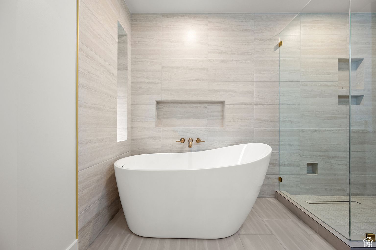This is a modern primary bathroom featuring a freestanding white bathtub as the focal point. The walls are covered in light gray, horizontally laid tiles, complemented by a built-in niche above the tub and a glass-enclosed shower with matching tile and built-in shelves. The overall impression is clean, luxurious, and spa-like.