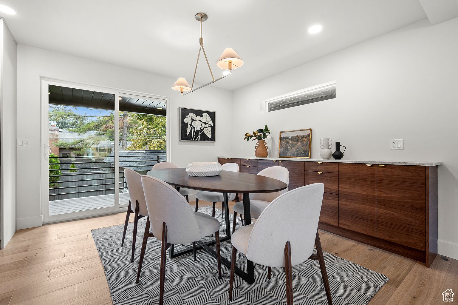 This is an interior shot of a dining room featuring a dark wood oval table surrounded by six light-colored upholstered chairs with dark wood legs. A modern chandelier hangs above the table, and a dark wood credenza with a light countertop sits against the wall, adorned with decorative items. A sliding glass door provides natural light and access to an outdoor area, while a geometric patterned rug anchors the space.