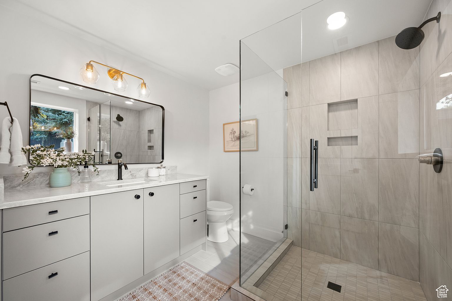 This is a well-lit primary bathroom featuring a modern design. The vanity has gray cabinets, a white countertop, and a black faucet, complemented by a black-framed mirror and a gold-toned light fixture. A glass-enclosed shower with beige tile and black hardware is visible, along with a toilet and a neutral-toned rug, creating a clean and contemporary space.