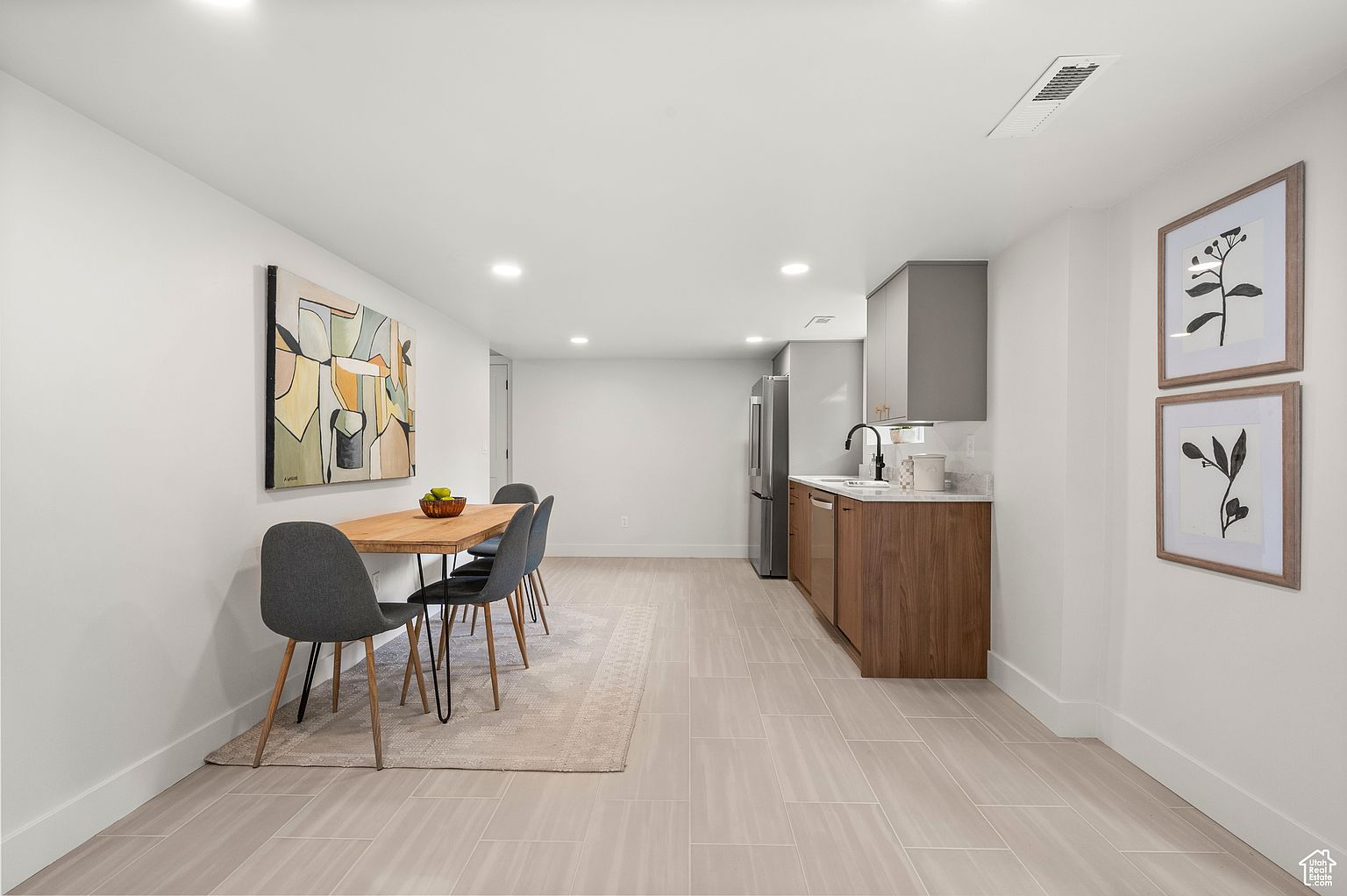 This interior shot showcases a dining area adjacent to a kitchen space. The dining area features a wooden table with modern gray chairs, complemented by an abstract painting on the wall and a neutral-toned rug underneath. The kitchen area includes sleek cabinetry and stainless steel appliances, creating a cohesive and modern living space.