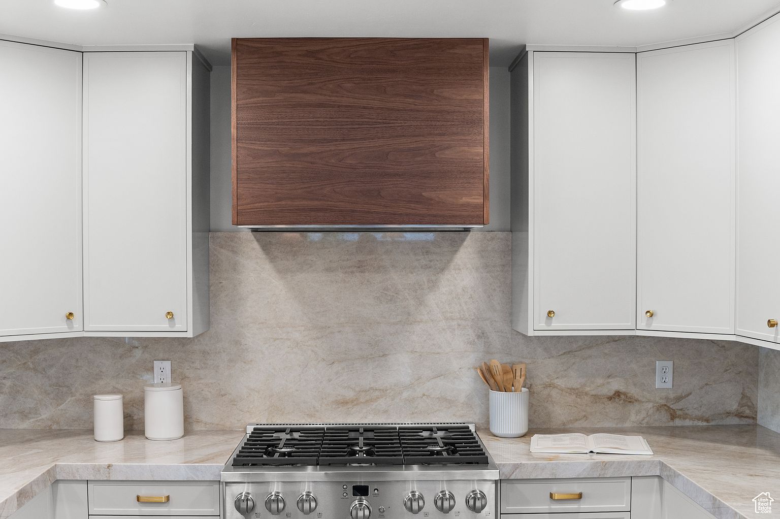 This is a well-lit kitchen featuring white cabinets with gold hardware and a contrasting wood range hood. The countertops and backsplash are a light marble, complementing the stainless steel range. The kitchen presents a clean and modern aesthetic.