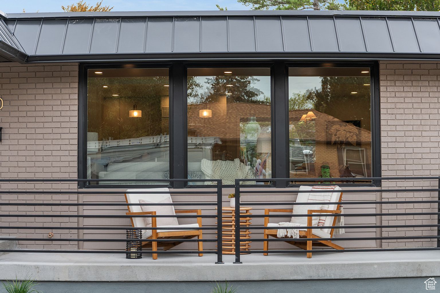 This image showcases a charming outdoor balcony or patio area. Two wooden chairs with light cushions flank a small, decorative table, creating an inviting seating arrangement. A black metal railing provides a modern contrast to the brick facade of the building, and large windows reflect the surrounding environment, adding depth to the scene.