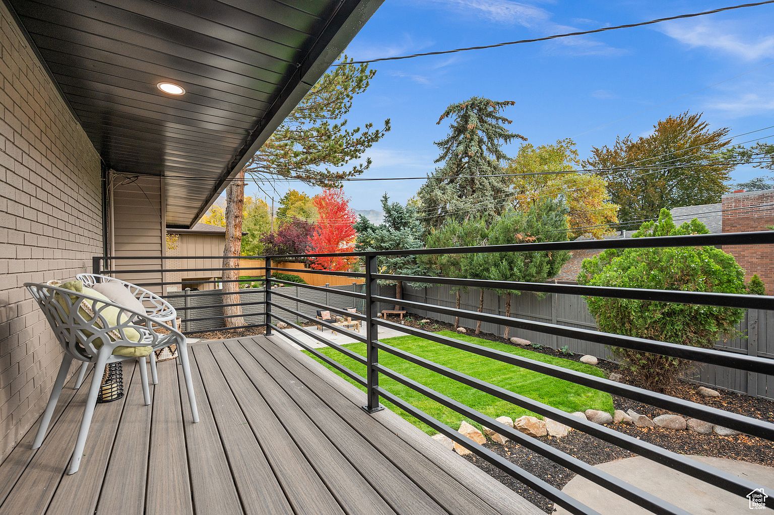 This image showcases a modern deck or balcony overlooking a well-maintained backyard. The deck features composite decking, a sleek black railing, and a stylish chair with cushions. The backyard includes a lush green lawn, landscaping with rocks and mulch, and a seating area with a fire pit, creating an inviting outdoor living space.