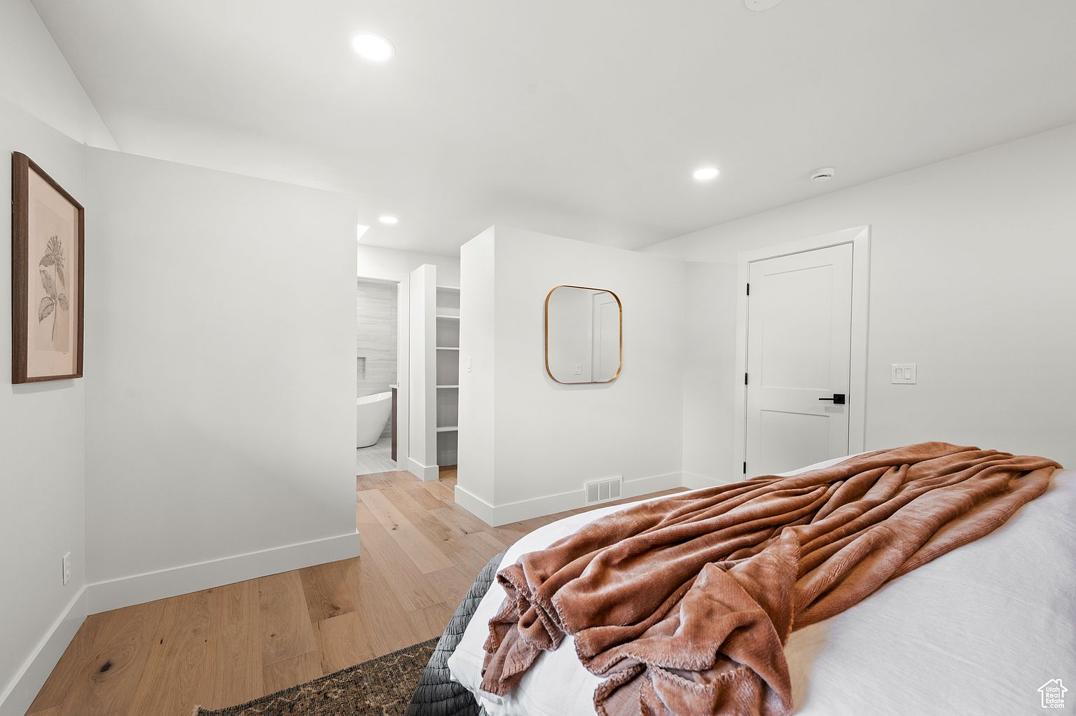 This is a bright and airy primary bedroom featuring hardwood floors, white walls, and a modern aesthetic. A large bed is draped with a plush, textured throw, and a minimalist mirror hangs on the wall. An open doorway leads to an ensuite bathroom, adding to the room's appeal.