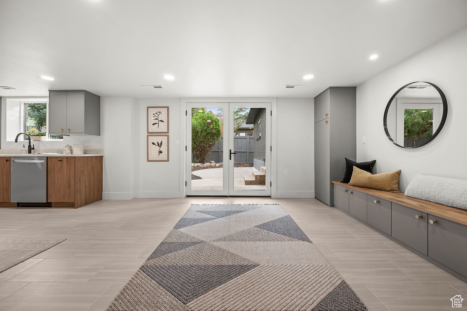 This is a well-lit basement area featuring a small kitchen area with modern cabinets and stainless steel appliances. The space includes a seating area with built-in storage and a decorative mirror. A large rug anchors the room, and glass doors lead to the outside, creating a bright and inviting atmosphere.