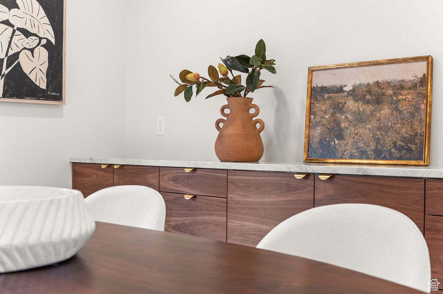 This interior shot showcases a dining room with a modern aesthetic. A dark wood table and white chairs are in the foreground, while a credenza with a marble top and wooden cabinets sits against the wall. Artwork and a vase with greenery adorn the credenza, adding a touch of sophistication to the space.