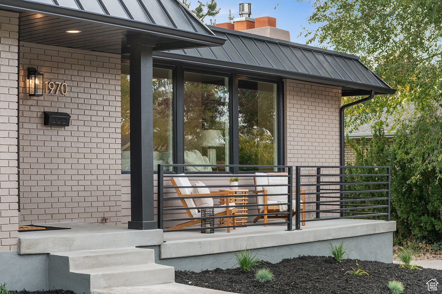This image showcases the entryway of a charming home, featuring a brick facade, a dark metal roof, and a covered porch. The porch is furnished with a cozy seating area, complete with wooden chairs and a small table, creating an inviting space. The well-maintained landscaping and modern railing add to the home's curb appeal.