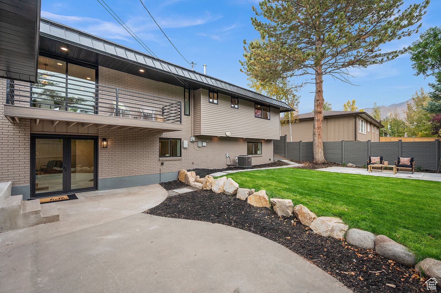 This is a rear view of a two-story house featuring a brick and siding exterior. A balcony with metal railings extends from the second floor, and a patio area is accessible through glass doors on the ground level. The backyard includes a well-maintained lawn, landscaping with rocks and mulch, and a privacy fence, creating an inviting outdoor space.
