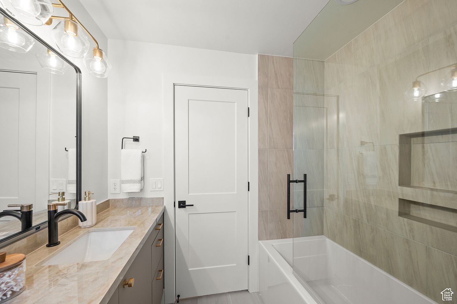This is a well-lit primary bathroom featuring a modern design. The vanity has a light brown countertop with gray cabinets and gold hardware. A large mirror with a gold-toned light fixture hangs above the sink. The bathroom also includes a bathtub with a glass enclosure and tiled walls, creating a spa-like atmosphere.