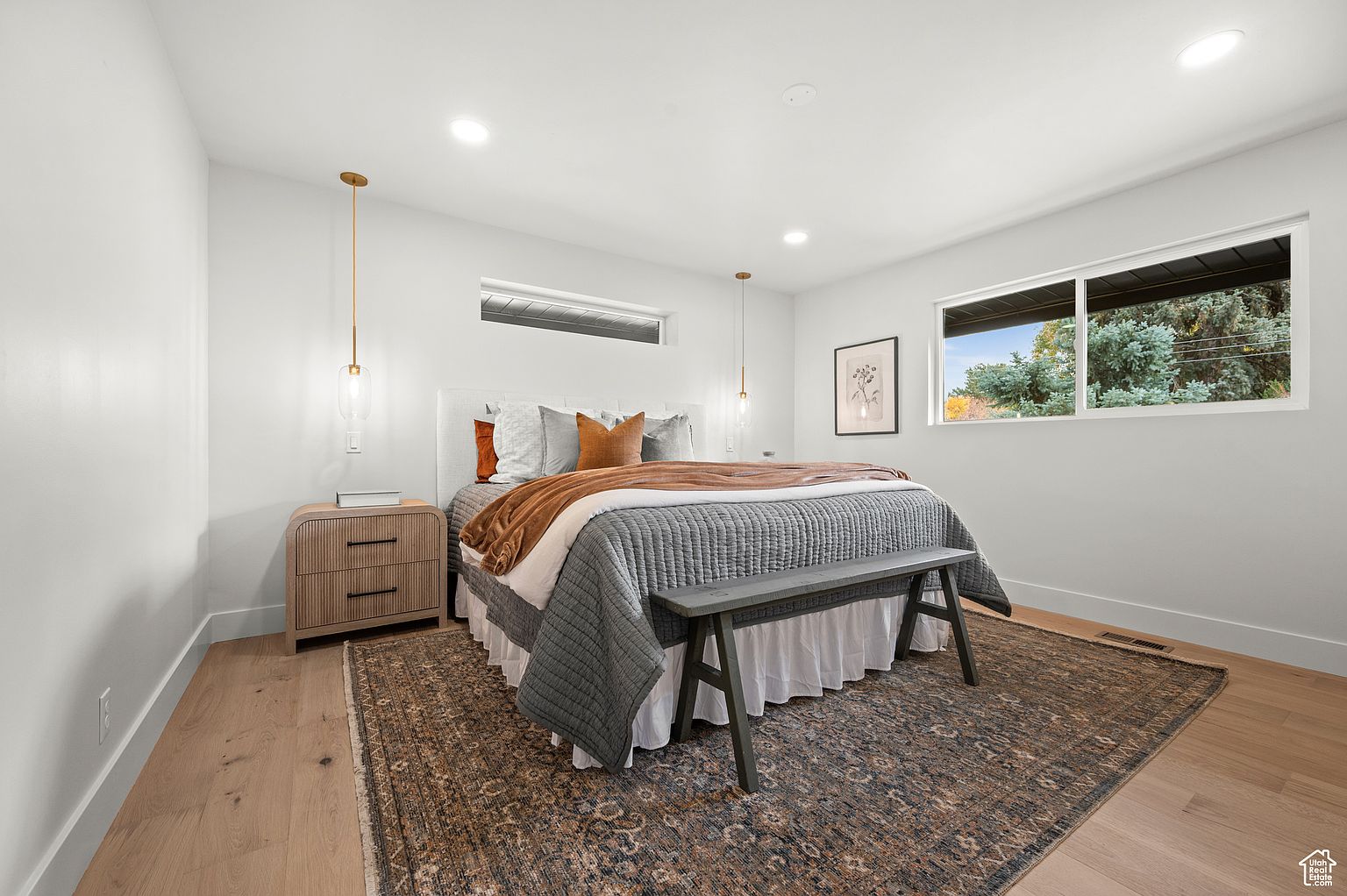 This is a bright and airy primary bedroom featuring a bed with a gray quilted coverlet and a white upholstered headboard. The room is decorated in neutral tones with wood flooring and a patterned area rug. Natural light streams in from a window, and pendant lights flank the bed, creating a relaxing and inviting atmosphere.