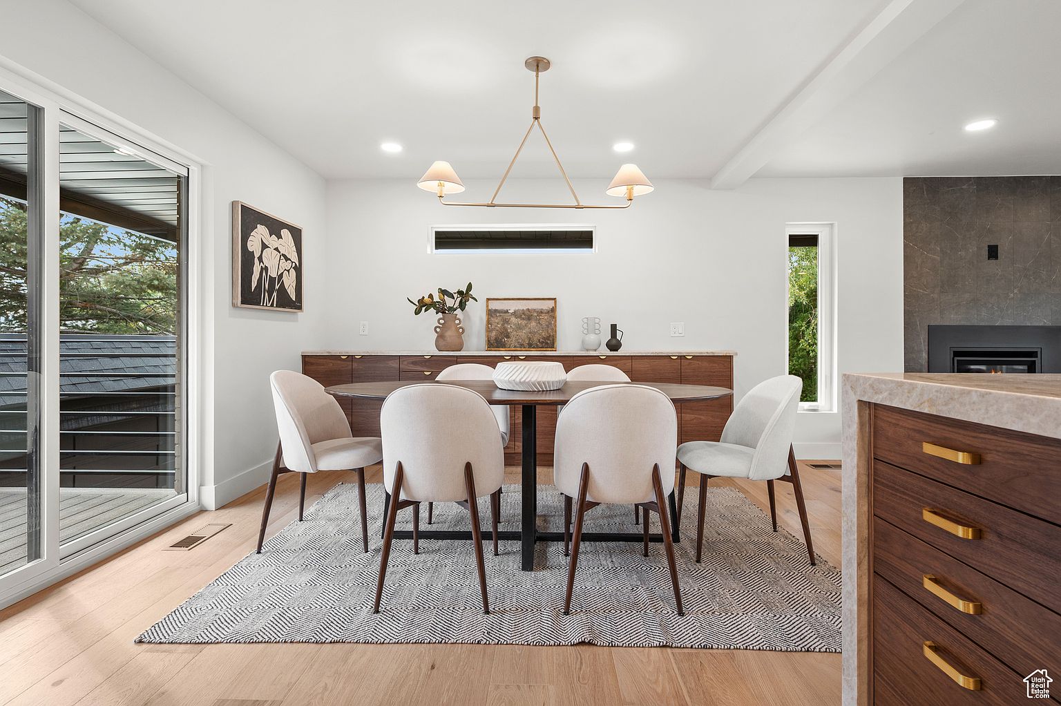 This is an interior shot of a modern dining room. The room features a wooden dining table surrounded by six upholstered chairs, set on a patterned rug. A long wooden credenza sits against the wall, adorned with decorative items, and a contemporary chandelier hangs above the table, creating a warm and inviting atmosphere.