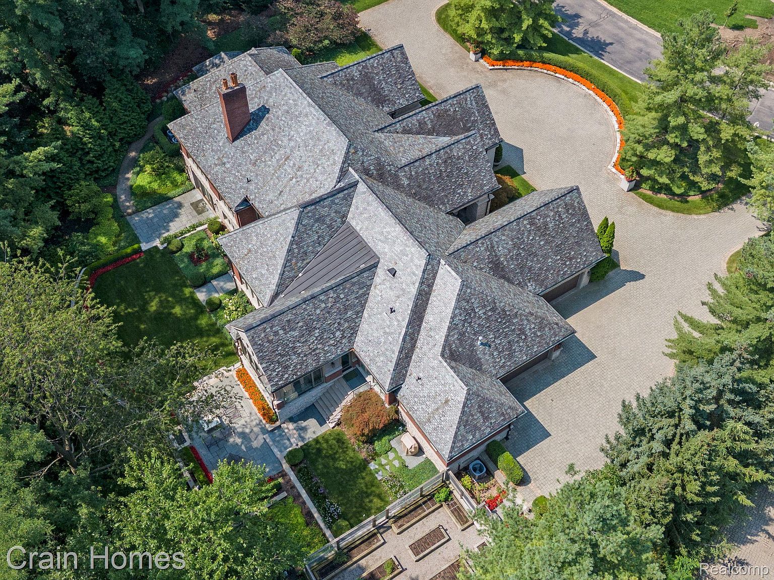 This aerial shot showcases a sprawling estate with a complex, multi-gabled roofline composed of gray shingles. The property features meticulously landscaped gardens, a circular driveway paved with interlocking stones, and mature trees providing privacy. The overall impression is one of luxury and seclusion.