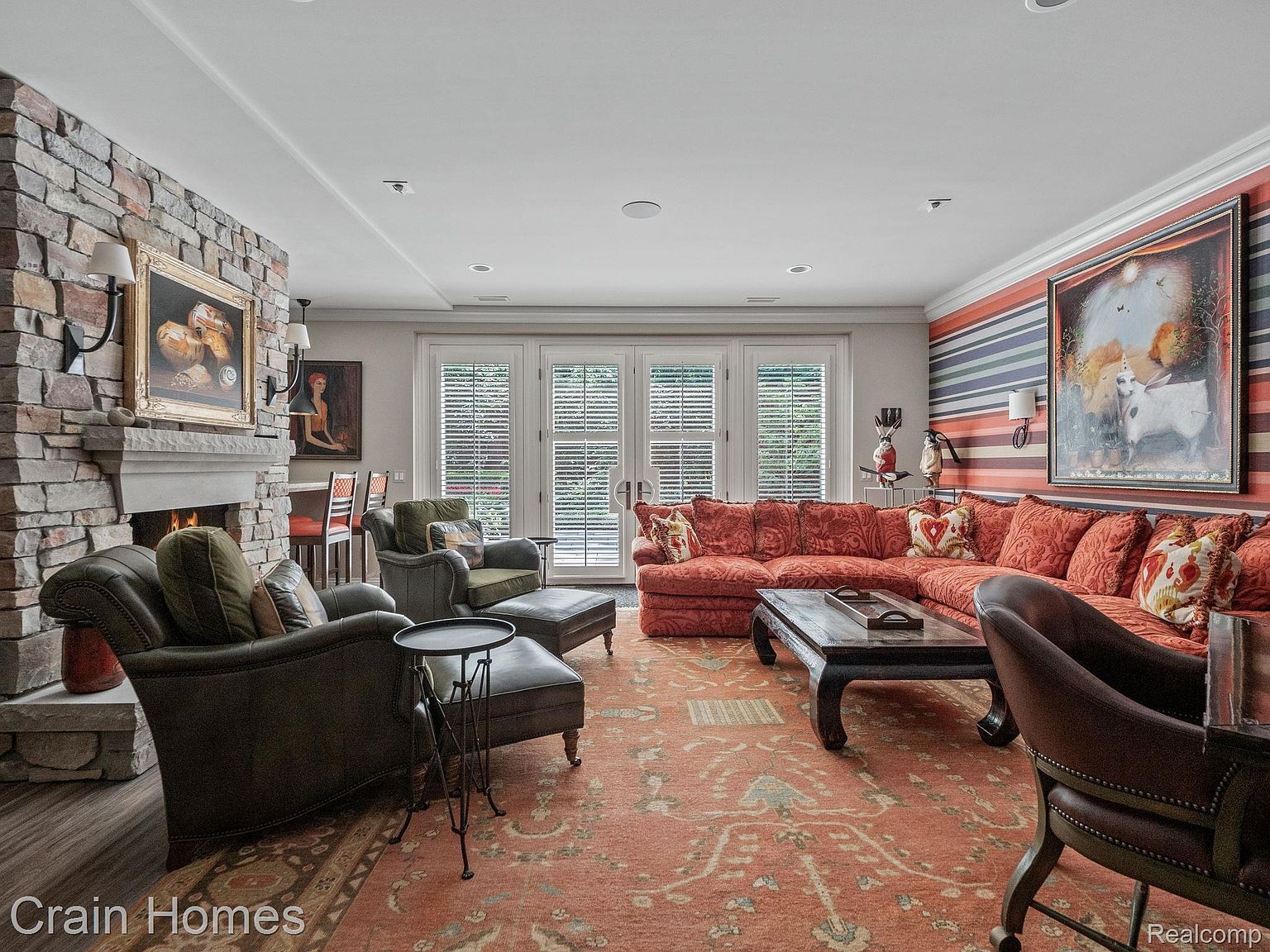 This is an interior shot of a living room featuring a stone fireplace, comfortable seating including a large sectional sofa and armchairs, and a patterned area rug. The room is well-lit with recessed lighting and sconces, and artwork adorns the walls, creating a warm and inviting atmosphere. French doors with blinds are visible in the background.
