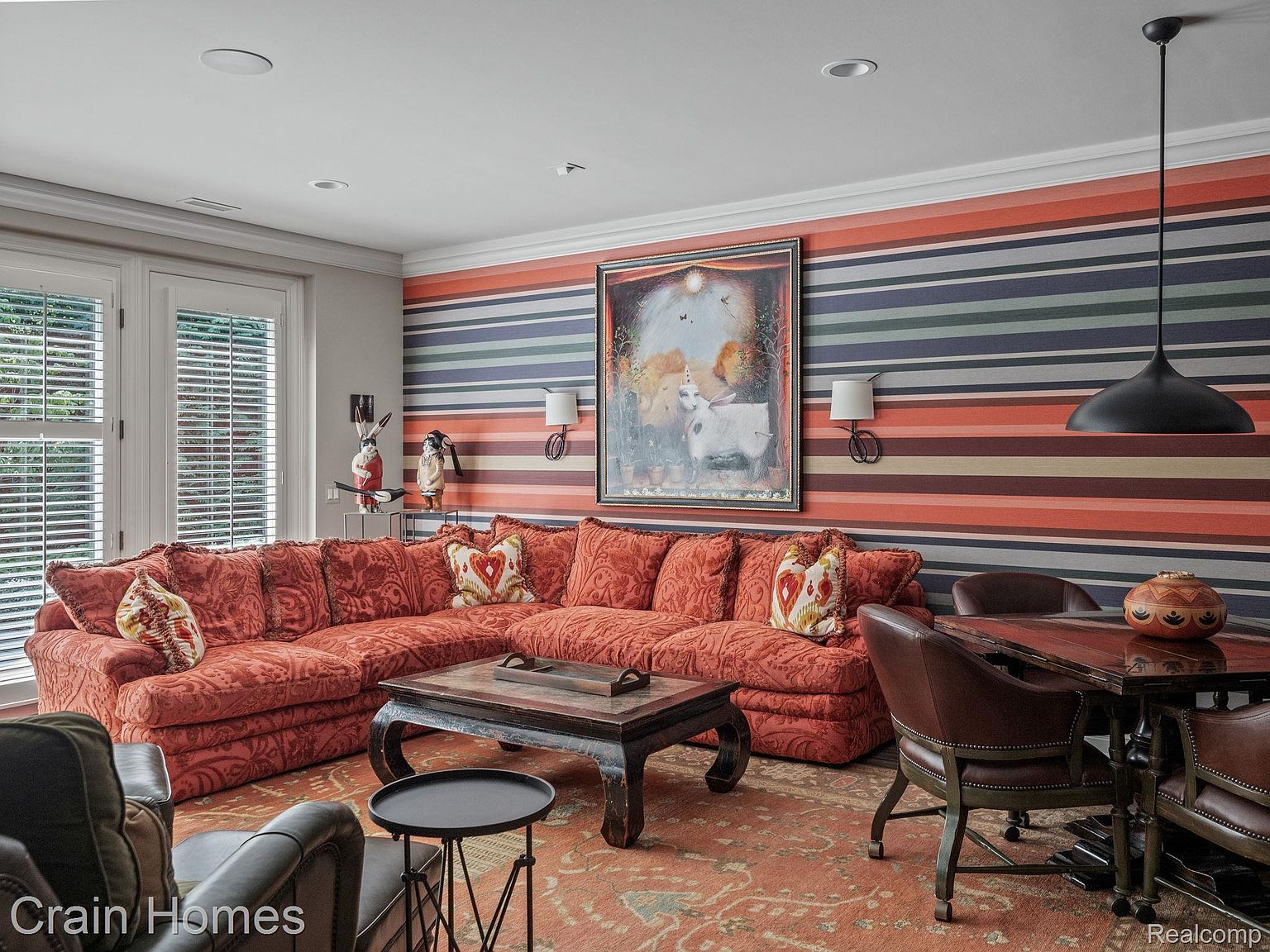 This interior shot showcases a cozy living room featuring a large, patterned orange sectional sofa, a dark wood coffee table, and a dining table with leather chairs. The room is decorated with a striped wallpaper, artwork, and unique decorative objects, creating a warm and inviting atmosphere. The perspective is from a medium distance, capturing the entire seating area and dining space.