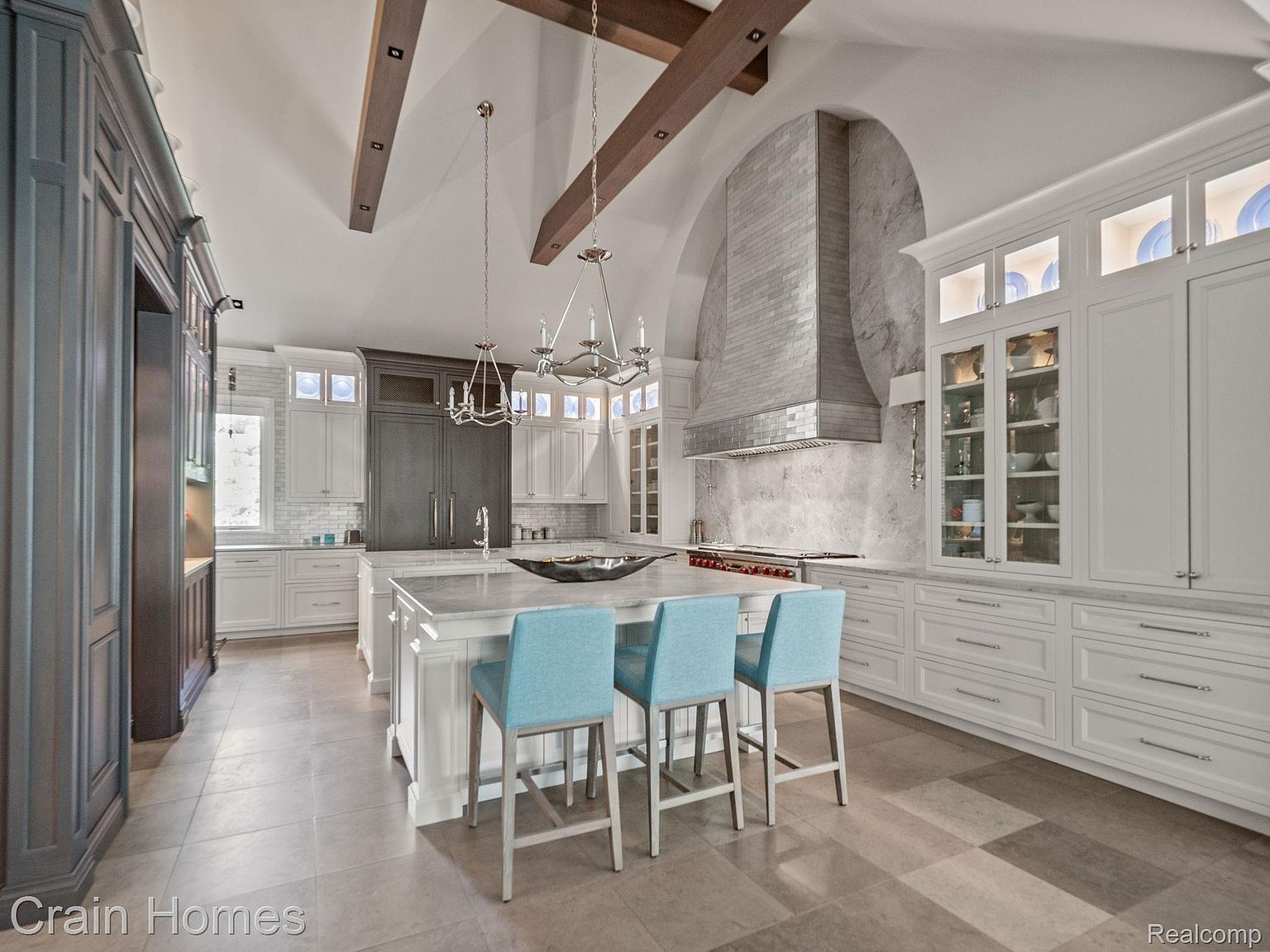 This is a spacious, high-end kitchen featuring custom cabinetry in both white and dark gray, complemented by a large central island with seating. The kitchen is equipped with stainless steel appliances and a statement range hood, while the design incorporates wooden beams and elegant chandeliers, creating a luxurious and inviting atmosphere. The flooring appears to be large format tile.