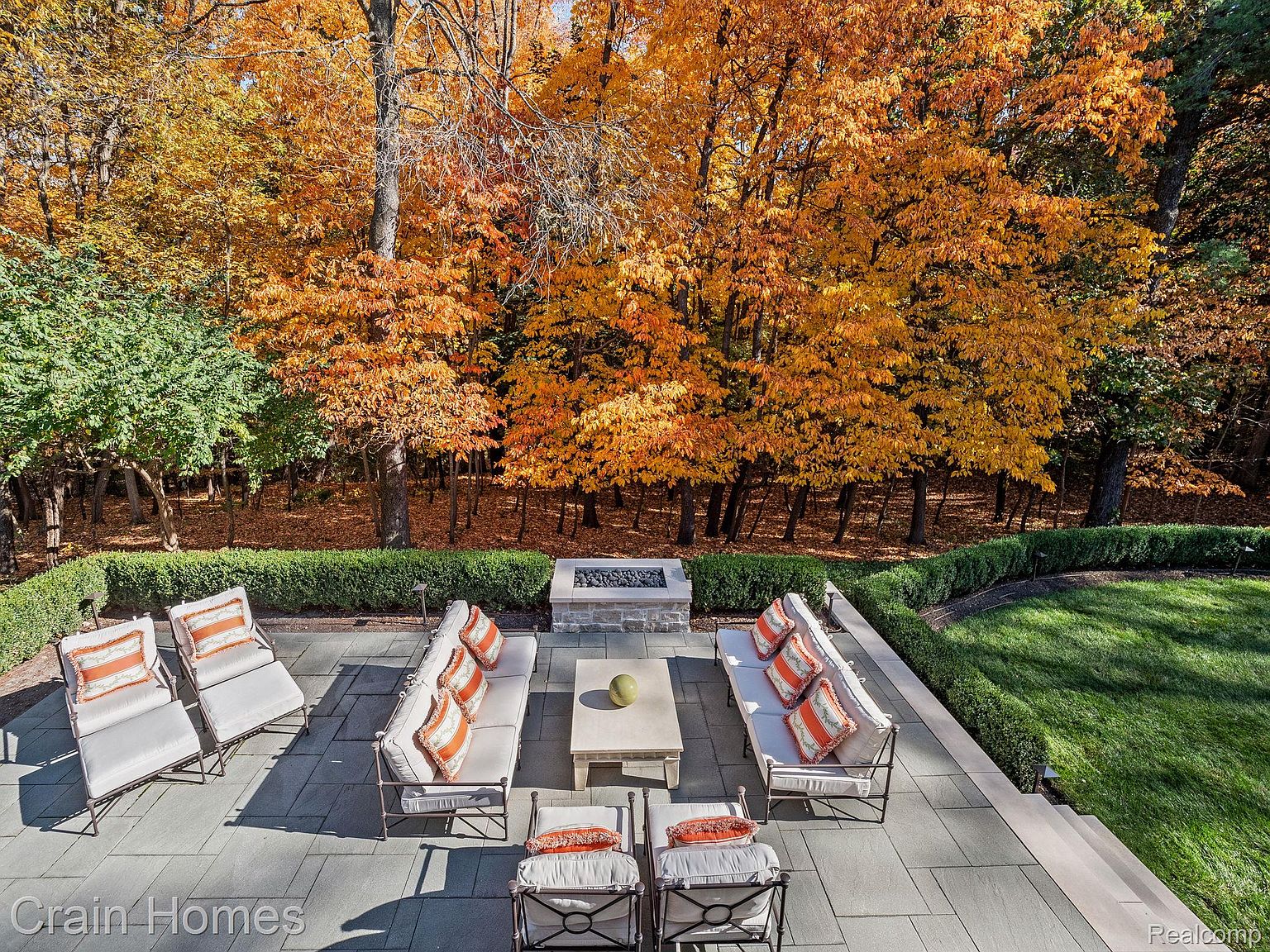 This is an aerial view of a beautifully designed outdoor patio area, perfect for relaxation and entertaining. The patio features comfortable seating arrangements with plush cushions and decorative pillows, centered around a modern fire pit and coffee table. The surrounding landscape showcases vibrant fall foliage, creating a serene and inviting atmosphere.