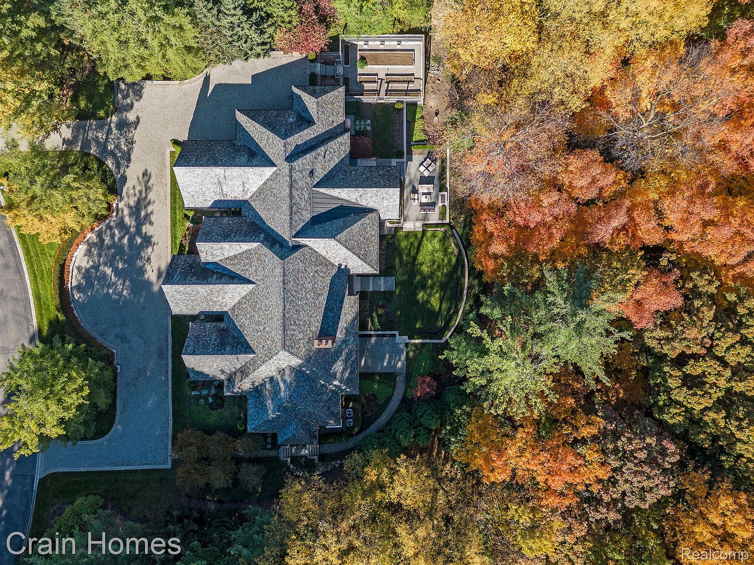 This aerial view showcases a large, luxurious home with a complex roofline and meticulously landscaped grounds. The property features a stone driveway, multiple patios, and a well-maintained lawn, all surrounded by mature trees displaying vibrant fall colors. The overall impression is one of grandeur and privacy.