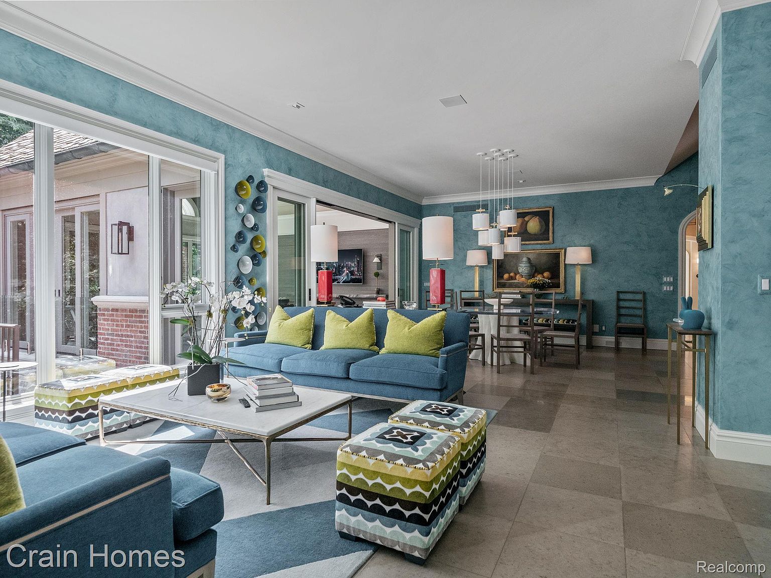 This is an interior shot of a living room and dining area featuring a blue color scheme. The living room has a blue sofa with yellow pillows, a patterned rug, and a marble-topped coffee table. The dining area includes a table with chairs and a painting on the wall, with the overall impression being stylish and coordinated.