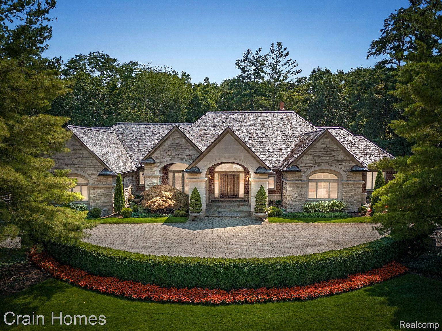 This is a front exterior view of a luxurious stone house with a well-manicured lawn and landscaping. The house features a grand entrance with an arched doorway, a circular driveway, and a meticulously maintained garden with vibrant flowers. The overall impression is one of elegance and high-end living.