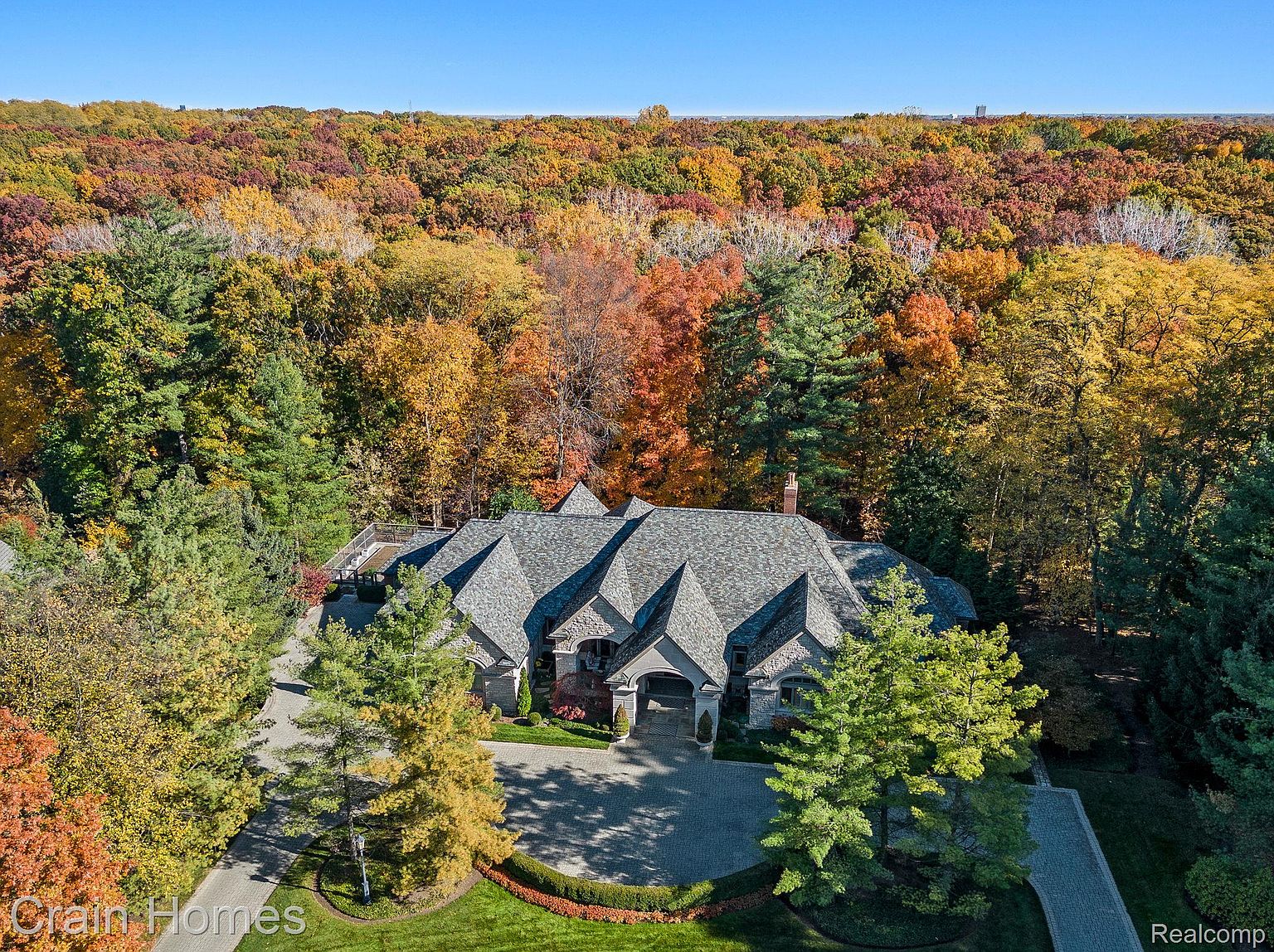 This aerial view showcases a grand estate nestled amidst vibrant fall foliage. The home features a complex roofline with multiple gables, a stone facade, and a circular driveway leading to a porte-cochère. Lush landscaping surrounds the property, enhancing its curb appeal and creating a sense of privacy and luxury.