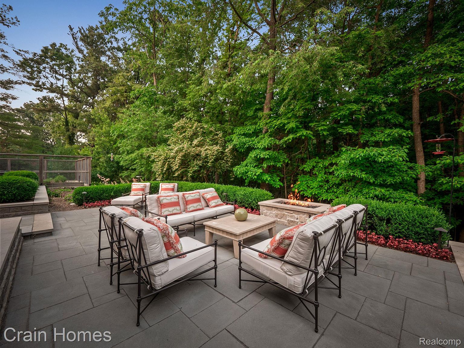 This is an inviting outdoor patio area featuring comfortable seating arranged around a stone fire pit. The patio is paved with large gray stones and surrounded by lush greenery, creating a private and serene atmosphere. The furniture includes cushioned chairs and a sofa with decorative pillows, perfect for relaxing and entertaining.
