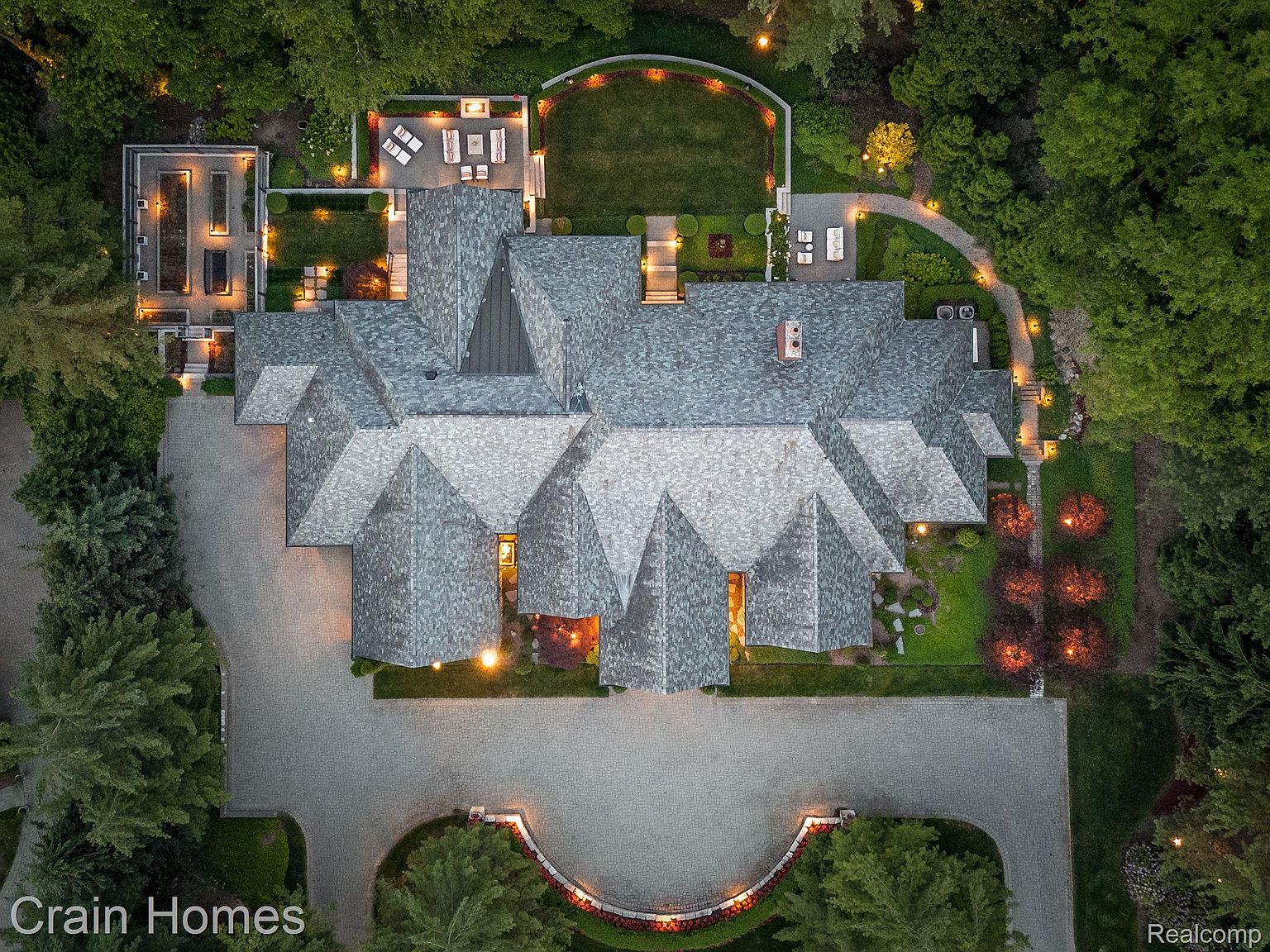 This aerial view showcases a sprawling estate with a complex, multi-gabled roofline and meticulously landscaped grounds. The property features a large driveway, manicured lawns, and strategically placed lighting that highlights architectural details and garden features. The overall impression is one of luxury, privacy, and sophisticated design.