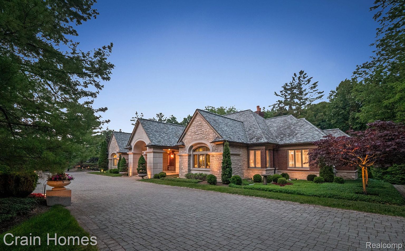 This is a front exterior view of a luxurious stone house with a well-manicured lawn and a brick driveway. The house features multiple gabled roofs, arched windows, and elegant landscaping. The overall impression is one of sophistication and curb appeal.