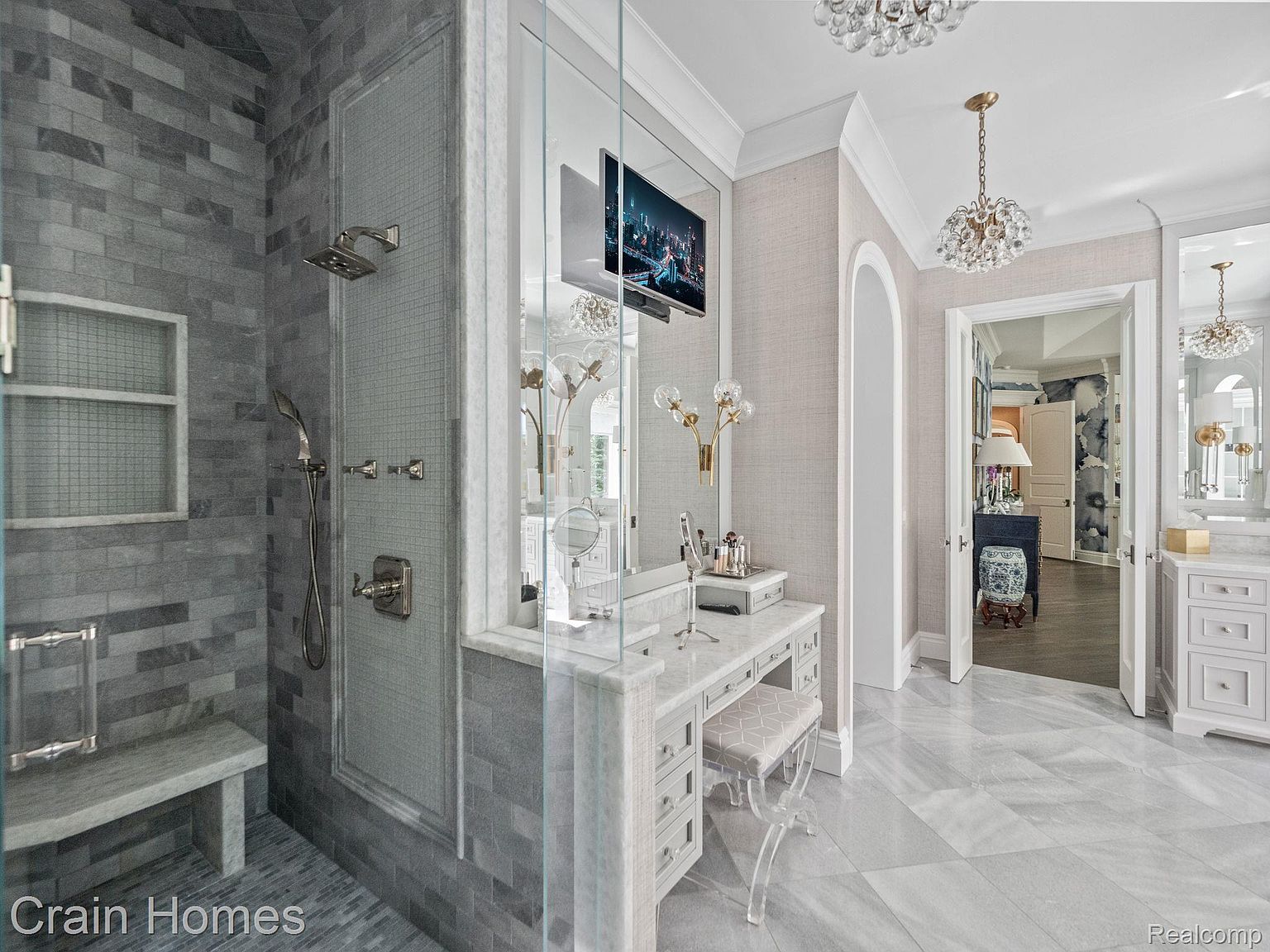 This is a luxurious primary bathroom featuring a walk-in shower with gray tiling and a built-in bench. A vanity area with a television and decorative lighting is adjacent to the shower, and the space is illuminated by crystal chandeliers. The flooring is light-colored marble, contributing to the bathroom's elegant and spa-like atmosphere.