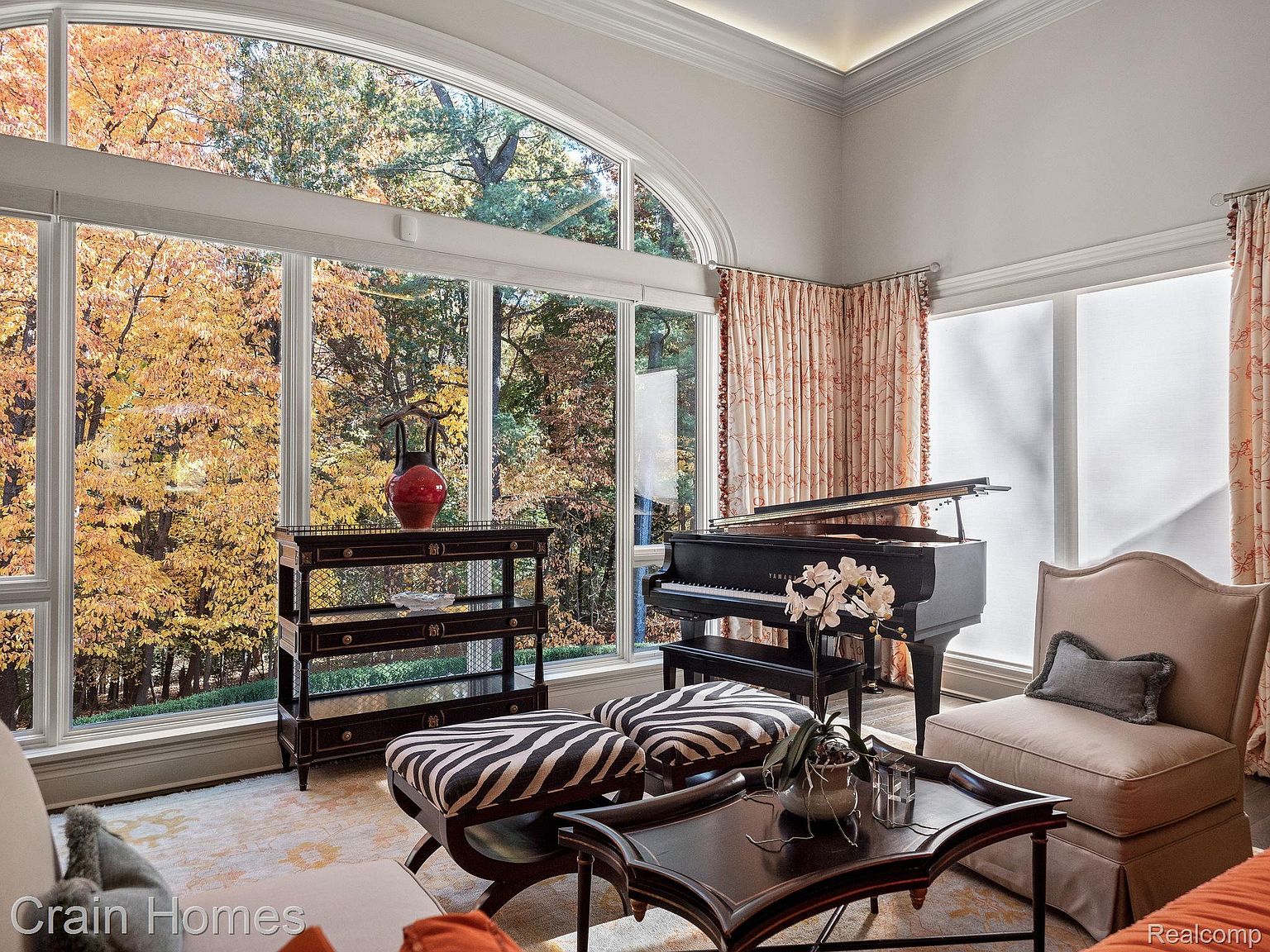 This is an interior shot of a luxurious living room featuring a grand piano, zebra-striped ottomans, and a dark wood console table. A large arched window provides a view of colorful autumn trees, enhancing the room's elegant and inviting atmosphere. The room is well-lit and decorated with tasteful furniture and accessories.