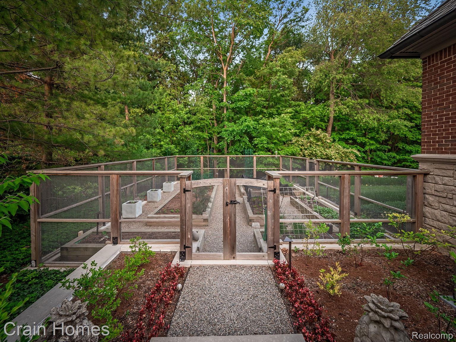 This image showcases a meticulously designed garden area, featuring raised garden beds enclosed by a wooden frame and wire mesh. A gravel pathway leads to the garden entrance, flanked by carefully arranged landscaping with colorful plants and decorative stone elements. The garden is set against a backdrop of lush trees, creating a serene and private outdoor space.