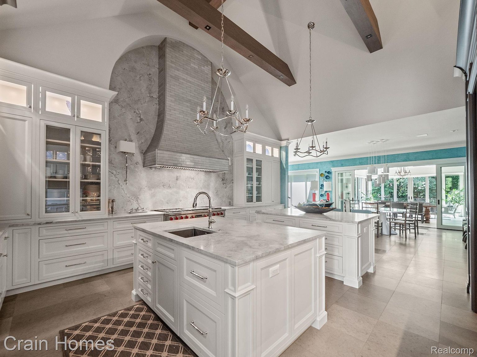 This is a bright and spacious kitchen featuring white cabinetry, marble countertops, and stainless steel appliances. The kitchen includes a large island with a sink and ample storage, as well as a decorative range hood. The open floor plan leads to a dining area with views of the outdoors, creating a seamless transition between spaces.