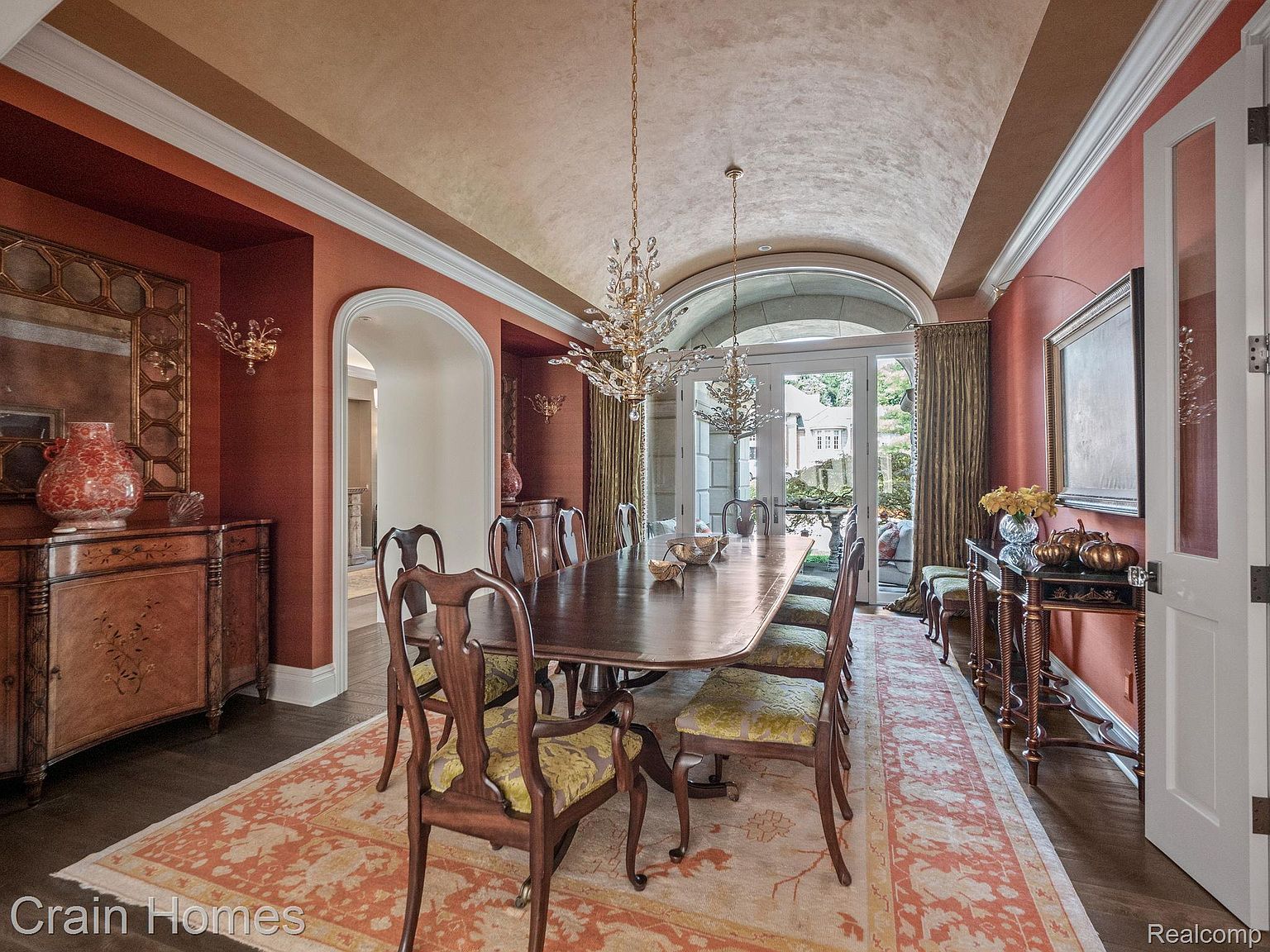 This is an interior shot of a formal dining room featuring a long wooden table surrounded by upholstered chairs. The room is decorated in warm tones with a textured wall finish, complemented by elegant chandeliers and a decorative rug. A doorway leads to another room, and a glass door provides natural light and a view to the outside.