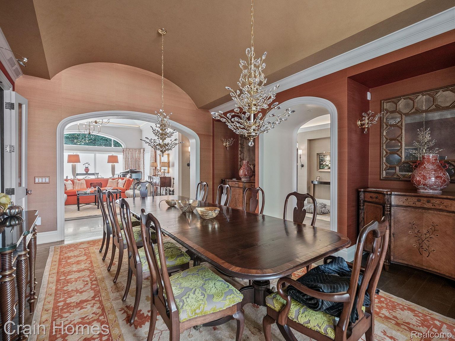 This is an elegant dining room featuring a long, dark wood table surrounded by ornate chairs with patterned cushions. Crystal chandeliers hang above the table, providing a luxurious ambiance. The room is decorated with rich, warm colors and textured wallpaper, creating a sophisticated and inviting space for formal dining.