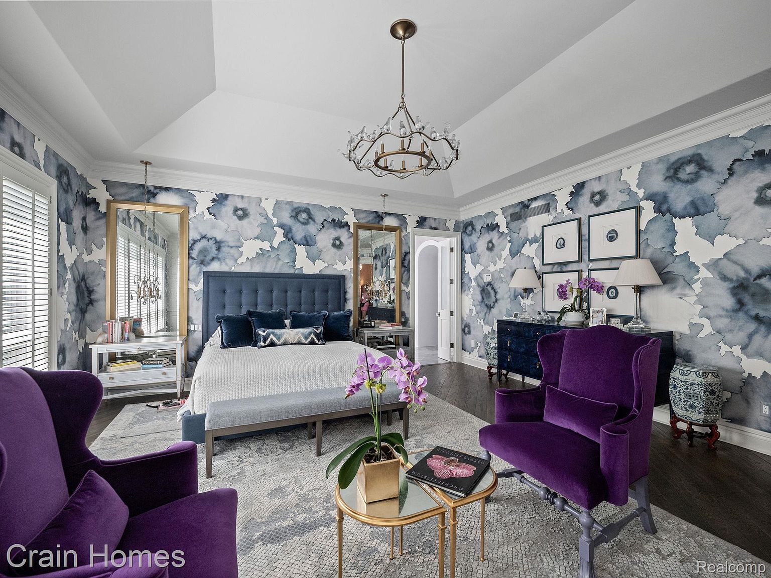 This is a luxurious primary bedroom featuring a king-size bed with a tufted headboard, complemented by a bench at the foot of the bed. The room is decorated with a striking floral wallpaper, accented by a modern chandelier and two elegant purple armchairs. The perspective is from the doorway, showcasing the room's depth and design.
