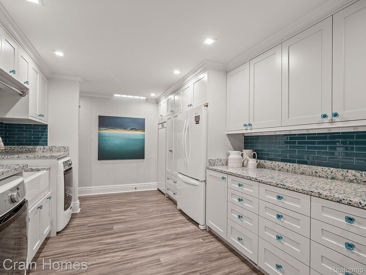 This is a bright and modern kitchen featuring white cabinetry with unique blue glass knobs and pulls, complemented by a speckled granite countertop and a blue subway tile backsplash. The kitchen is well-lit with recessed lighting and includes stainless steel appliances, creating a clean and inviting space. The flooring is a light wood-look plank, and a piece of artwork adds a pop of color to the adjacent wall.