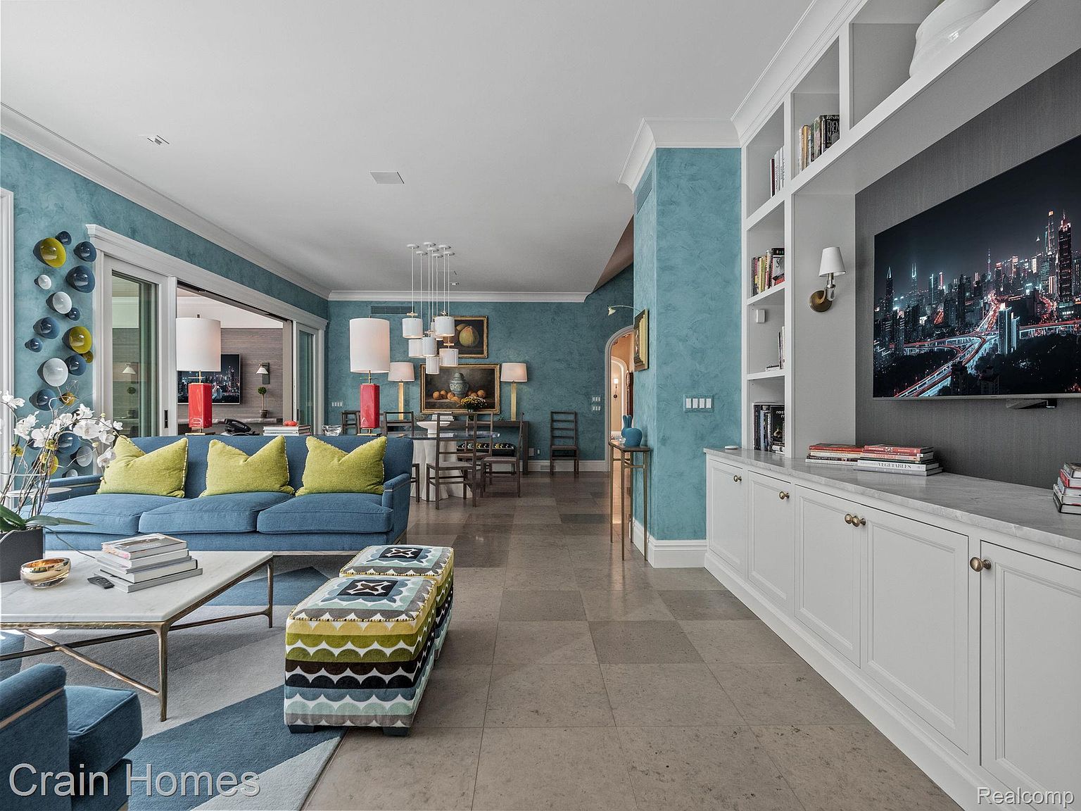 This is an interior shot of a luxurious living room featuring a blue sofa with yellow pillows, a patterned ottoman, and a marble-topped coffee table. The room has a sophisticated color palette with blue walls and neutral-toned flooring. A built-in shelving unit with a television adds a modern touch, while the open layout connects to the dining area, creating a spacious and inviting atmosphere.